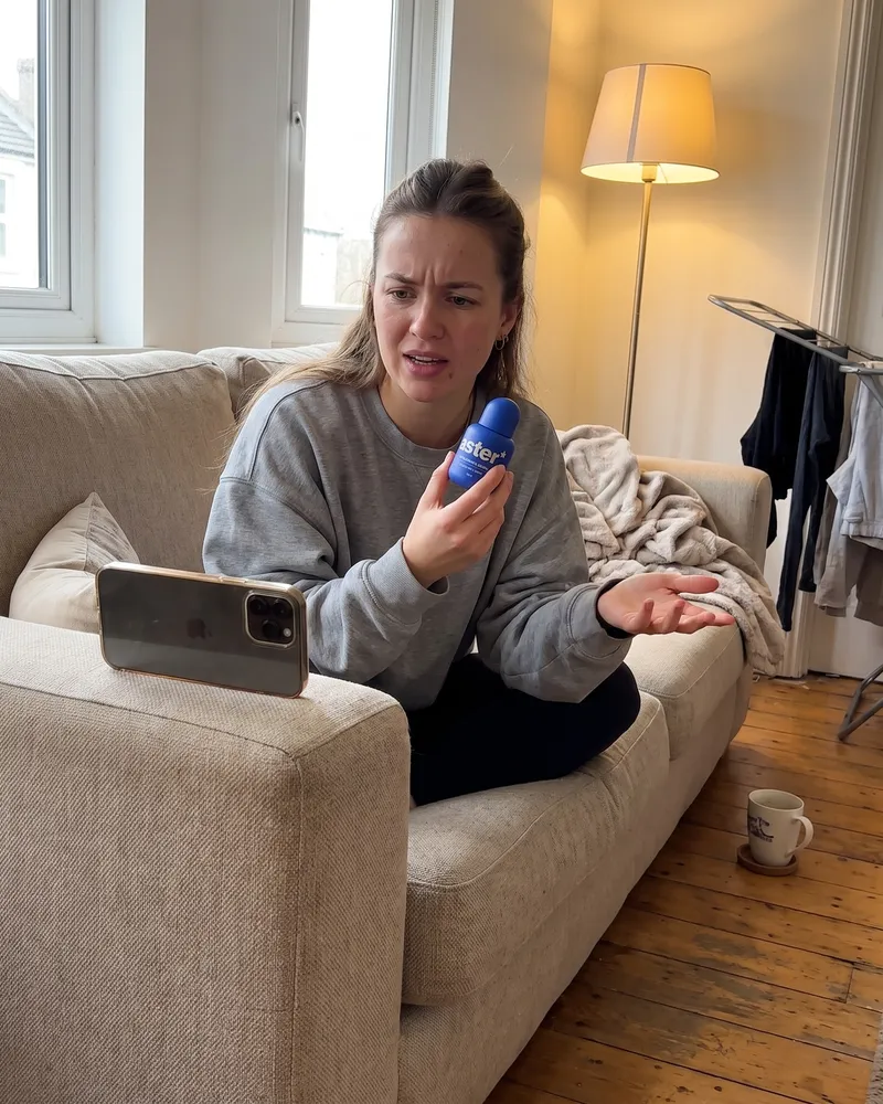 Example artwork for AI UGC review: A creator on her sofa filming herself on a propped-up phone, holding the bottle up